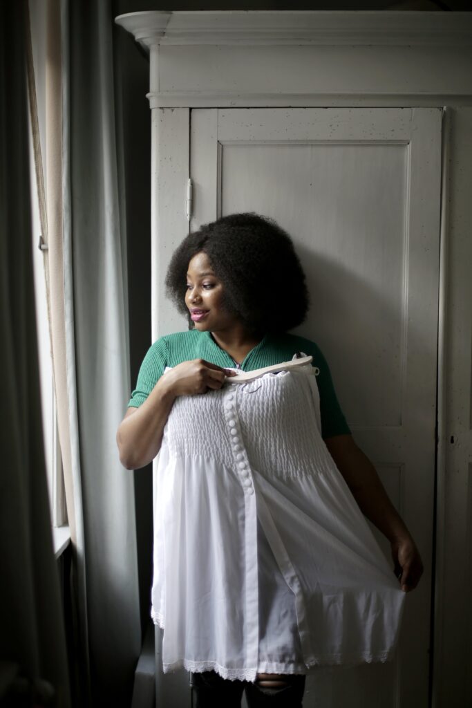 Embrace your femininity - Confident woman with afro hairstyle posing in front of a white door holding up a white dress