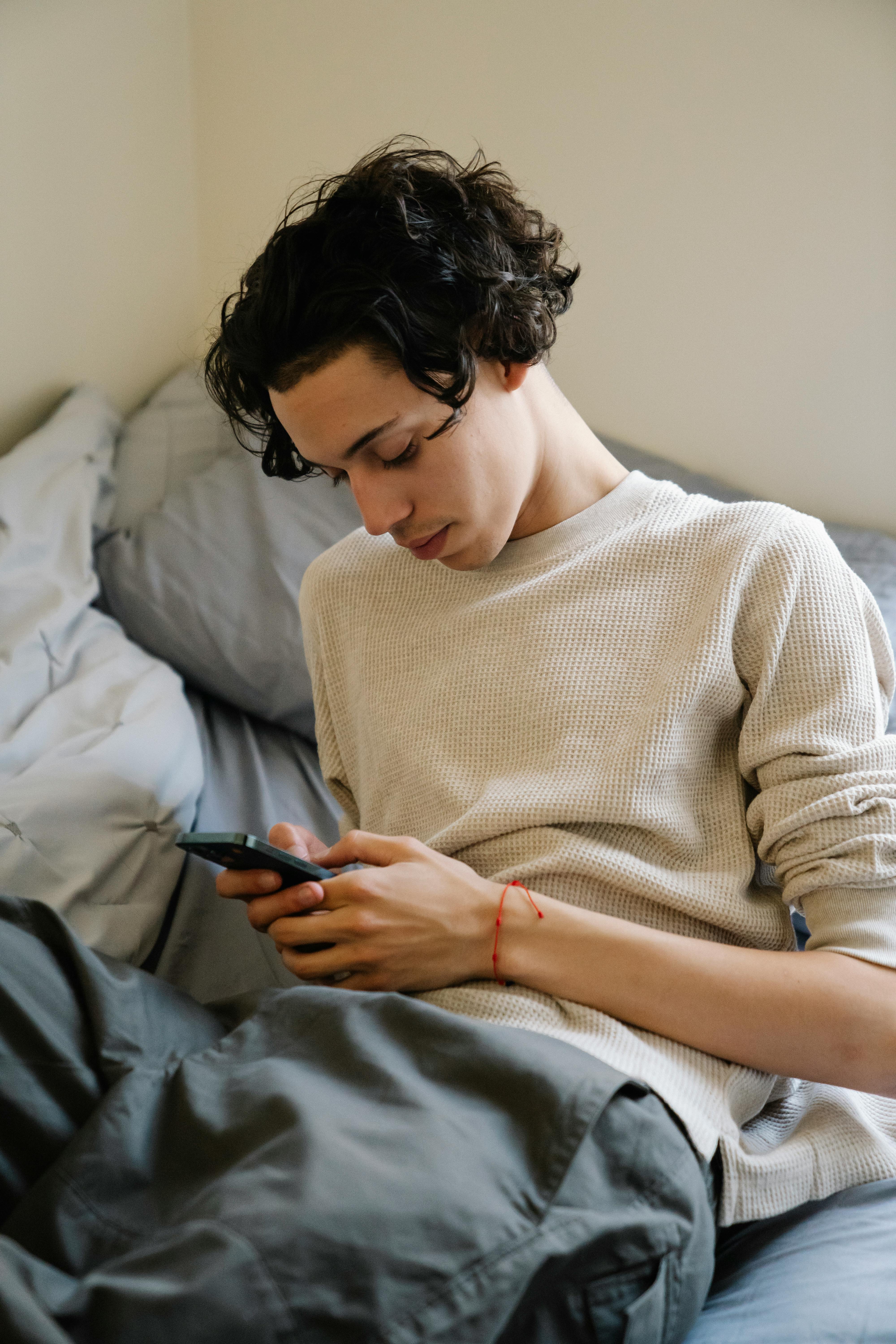 self care sunday - brown haired man lays on his bed scrolling on his phone, looking distressed