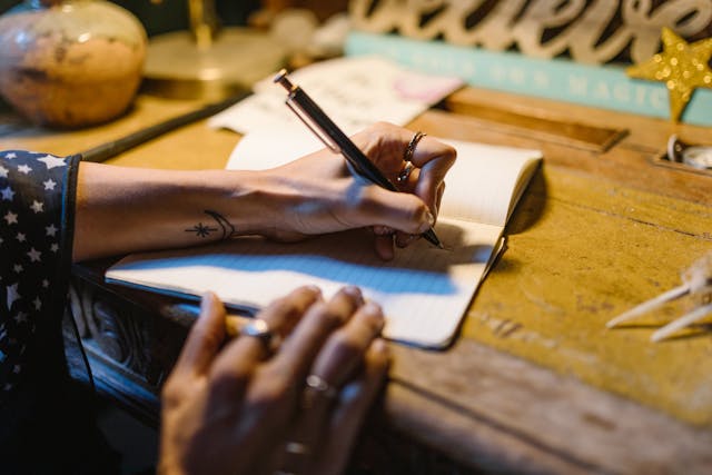 Types of journaling: person sitting at a desk with one hand writing in a notebook and the other resting on the edge of the table, holding the corner of the notebook